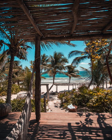 Wooden deck under a rustic palm-thatch pergola looking out through tropical gardens to a white sand beach