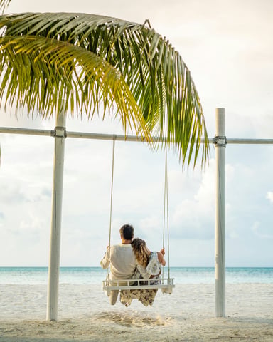 Couple sitting on a beach swing under a palm frond, facing the turquoise Caribbean sea