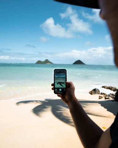 Person holding a phone on a white sand beach, photographing two small islands in turquoise water