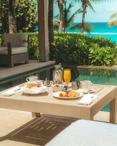 Breakfast table by a villa pool with fruit, croissants, and orange juice, turquoise sea in the background