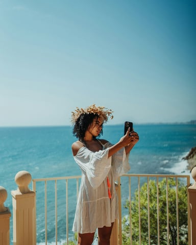 Woman in a white dress taking a photo from a clifftop balcony overlooking the blue Mediterranean