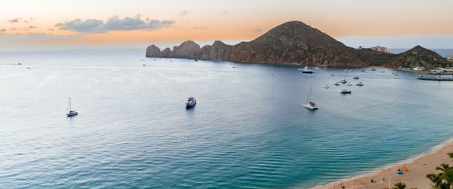 Elevated view of Cabo San Lucas bay at sunset, sailboats anchored in calm water with El Arco rock formation and mountains in the distance